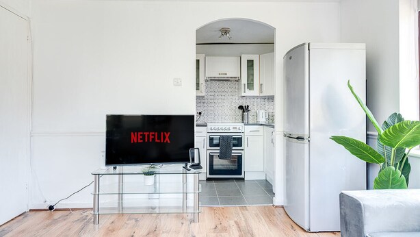 Ett modernt vardagsrum med en platt-TV, ett soffbord i glas, en vit soffa, en stor grön växt och ett kök i bakgrunden.