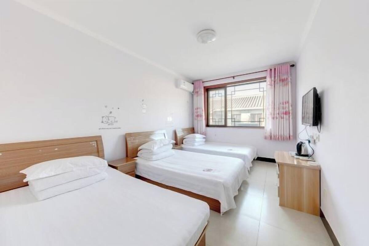 A hotel room with three beds, a wooden headboard, a small desk with a kettle, and a window with pink curtains.