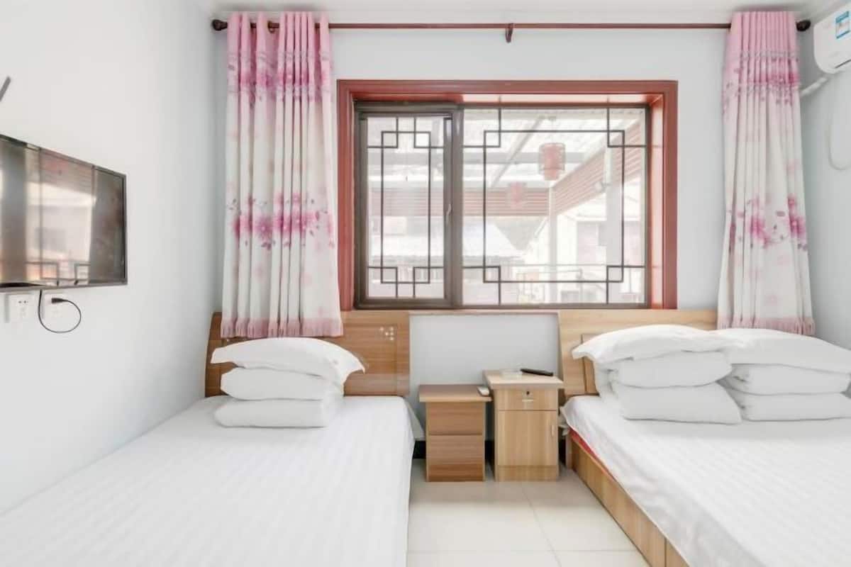 A bedroom with two beds, a TV, a window with curtains, and a small bedside table.