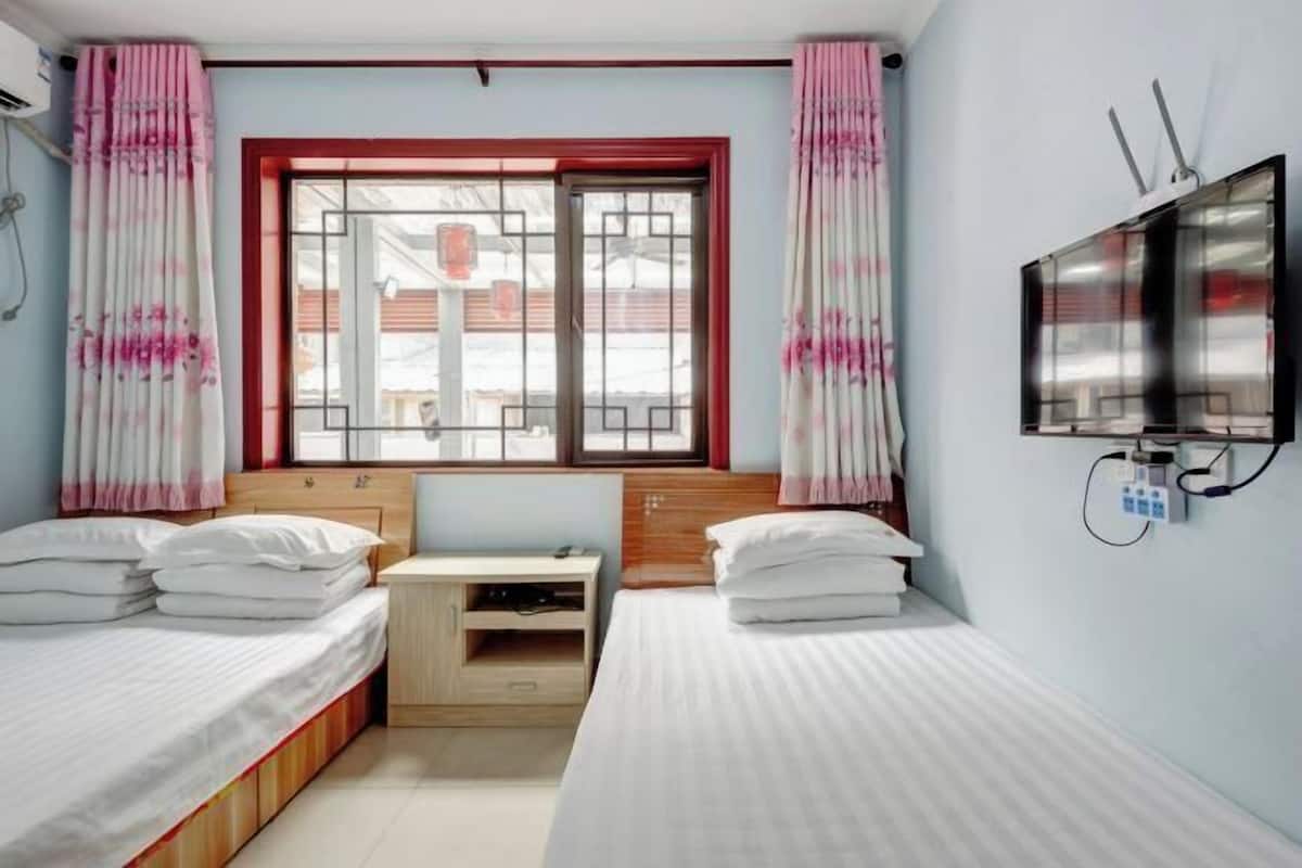 A bedroom with two single beds, a wooden desk, a TV mounted on the wall, and a window with a view of buildings.