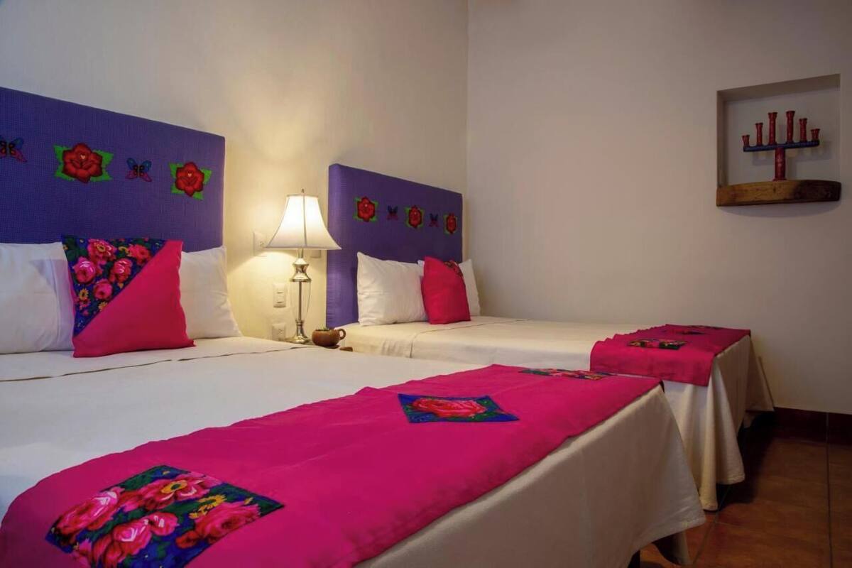 Hotellihuoneessa on kaksi sänkyä, joissa kummassakin on violetti pääty ja vaaleanpunainen tyyny. Sängyn vieressä on yöpöytä, jossa on lamppu, ja seinälle on kiinnitetty hylly, jossa on koriste-esineitä.