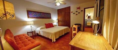 A hotel room with a bed, a chair, a desk, a ceiling fan, and a wall with artwork.