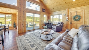 A spacious living room with a leather sofa, a coffee table, and a fireplace. There are large windows and a view of the outdoors. - Lakefront Divide Cabin w/ Game Room! (Divide)