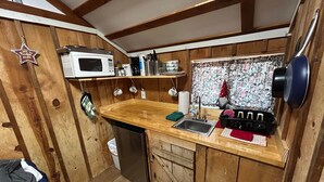 A compact kitchen with wooden walls, a microwave, a sink, and a window with a curtain. - Cabin #6 - Studio - No Fireplace (Payson)