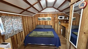 A room with wooden walls, a bed with blue bedding, a mounted picture, a microwave, and a small window. - Cabin #6 - Studio - No Fireplace (Payson)