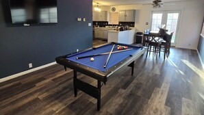 A pool table in a room with a ceiling fan, a TV mounted on the wall, and a kitchen area in the background. - Charming 4-bedroom house with beautiful backyard and mountain view. (Albuquerque)