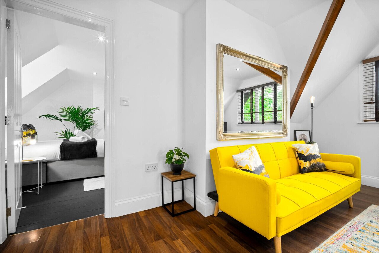 Um sofá amarelo vibrante com um espelho acima, uma pequena mesa de apoio de madeira com uma planta, e uma escada que leva a um andar superior.