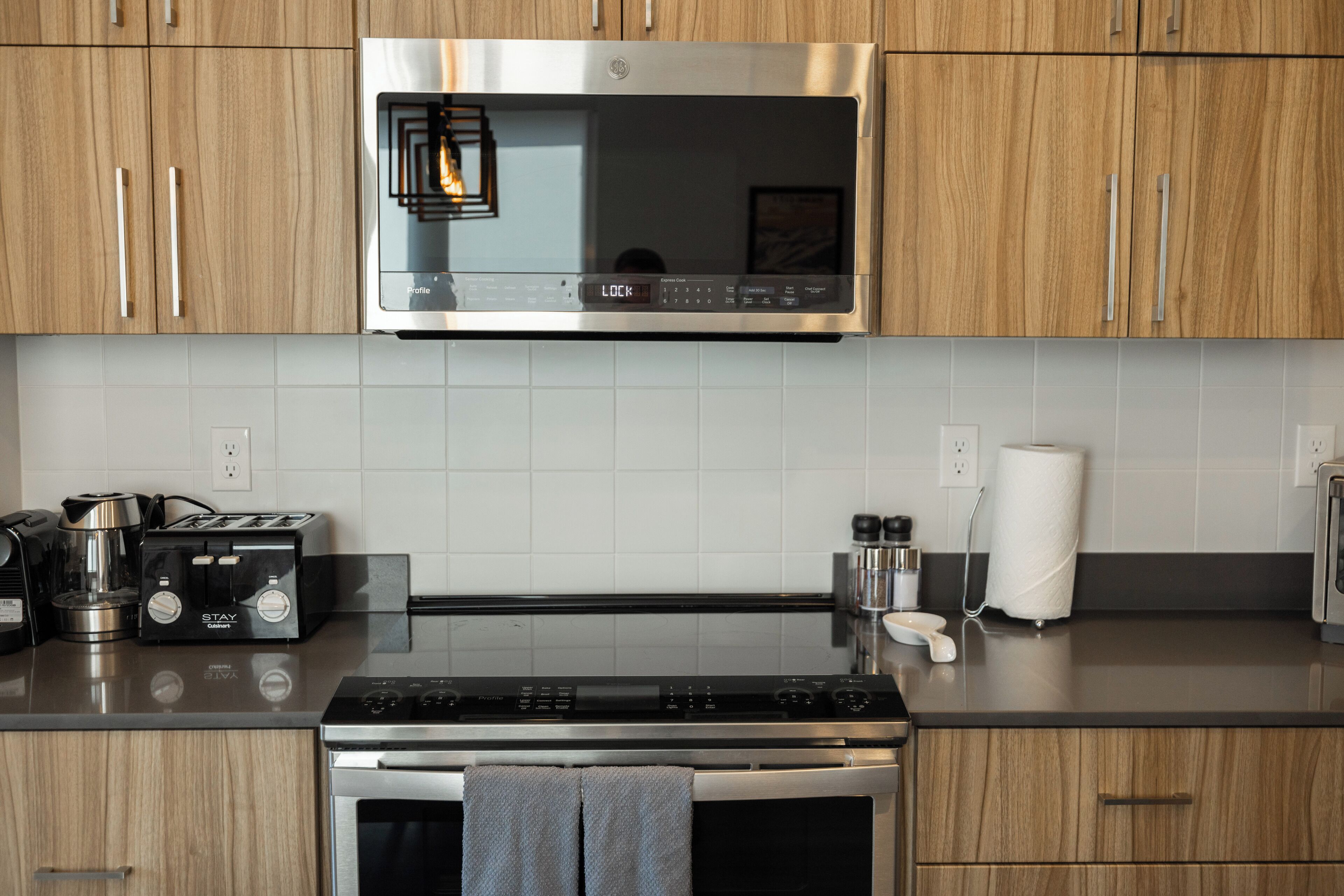 A modern kitchen with wooden cabinets, a stainless steel microwave, a built-in oven, a coffee maker, a toaster, and a kettle.