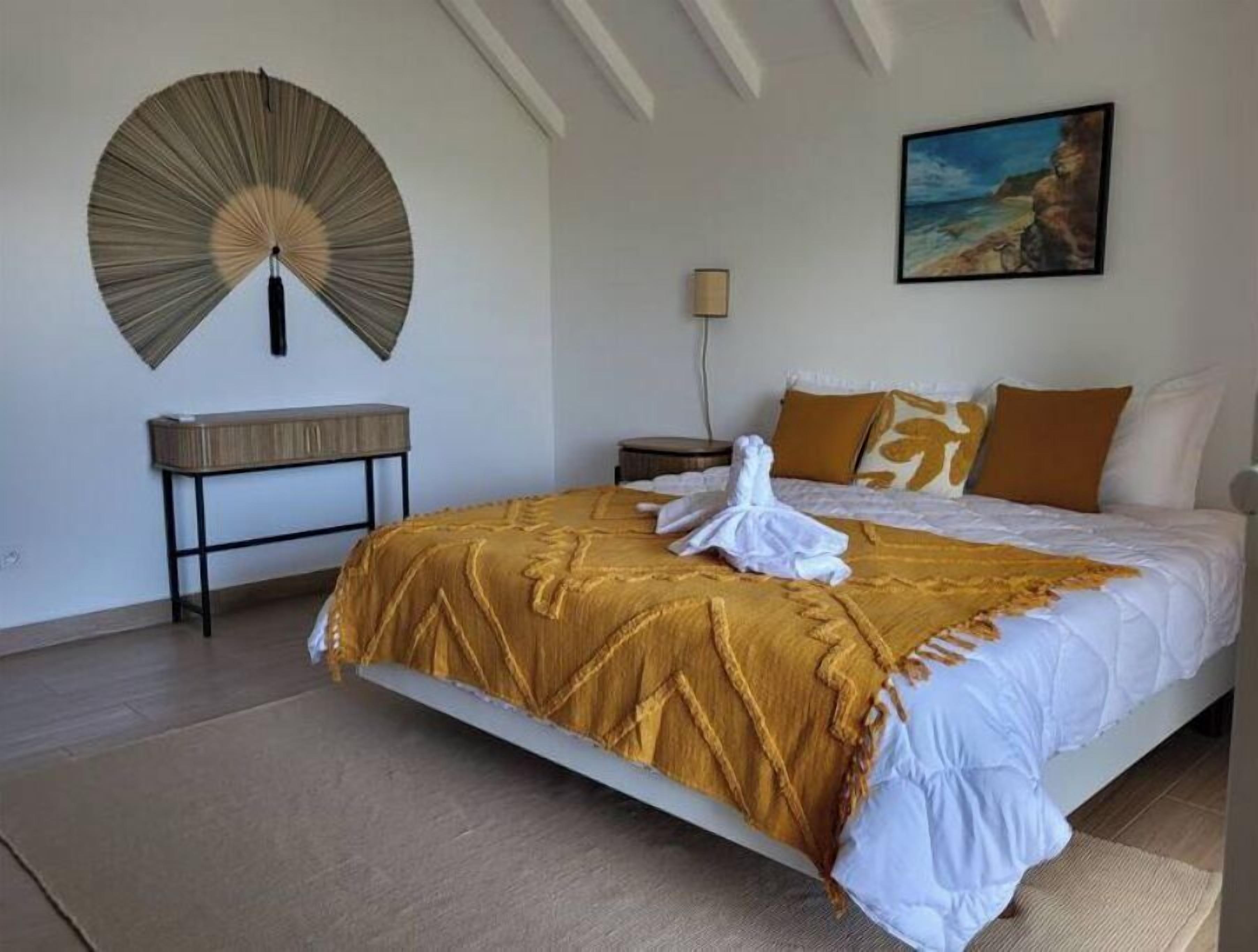 A bedroom with a large bed, a bedside table, a wall-mounted fan, a painting, and a lamp.