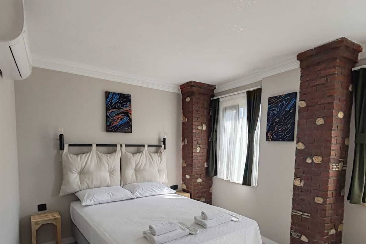 A bedroom with a bed, two pillows, a bedside table, a brick column, and artwork on the walls.