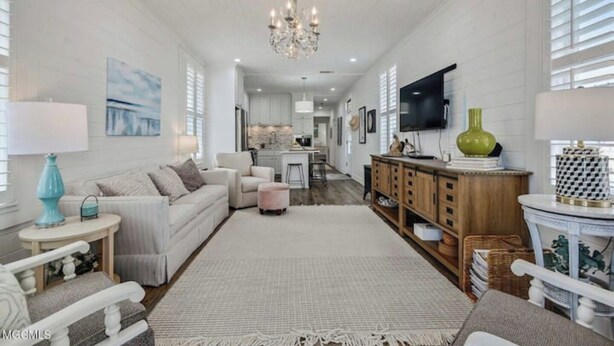A spacious living room with a neutral color palette, a chandelier, a television, and a kitchen in the background. - Beauty and The Bay (Bay St. Louis)