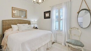 A bedroom with a large bed, a bedside table, a hanging mirror, and a window with curtains. - Beauty and The Bay (Bay St. Louis)