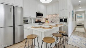 A modern kitchen with white cabinetry, stainless steel appliances, and a central island with stools. - Beauty and The Bay (Bay St. Louis)