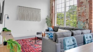 A spacious living room with a large gray sectional sofa, a wooden coffee table, a red patterned rug, and a ceiling fan. - Warm, Cozy 2BD | Pool | Gym | Central! (Greenville)