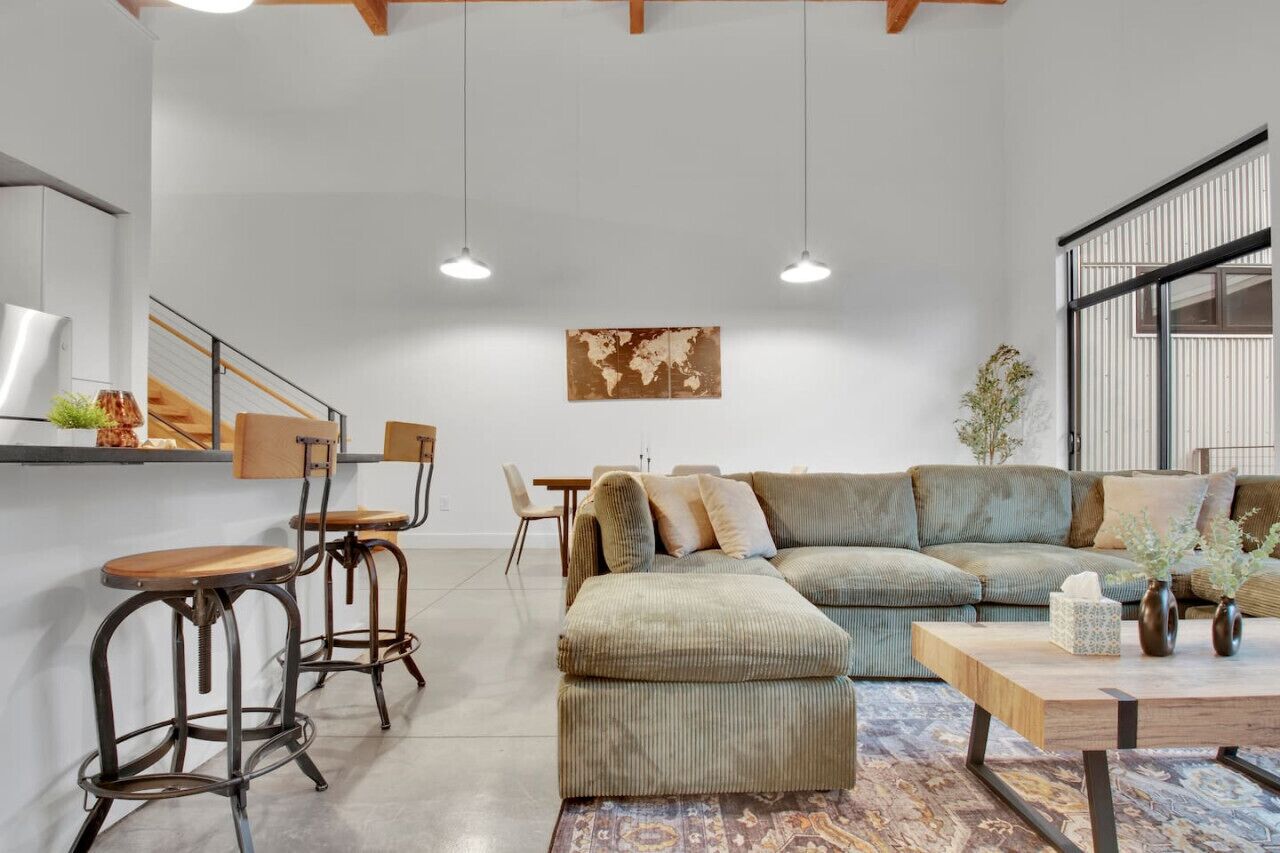 A modern living room with a green sofa, wooden coffee table, and metal bar stools. There is a world map on the wall and a staircase with wooden railings.