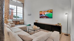 A modern living room with a large window, a sectional sofa, a round coffee table, and a flat-screen TV mounted on the wall. - Huge, Open Loft | Arts District | 5 mins to DT GVL (Greenville)