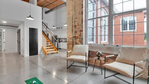 A spacious room with a high ceiling, exposed brick wall, and large windows. There are two white chairs and a small table with a globe. A staircase is visible in the background. - Huge, Open Loft | Arts District | 5 mins to DT GVL (Greenville)