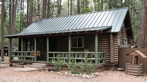A rustic log cabin with a metal roof, a porch with green railings, and a stone chimney. - Cabin #8 - Ranch House (Payson)