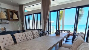 Un comedor con una gran mesa de madera y sillas acolchadas, ventanas amplias con vista al mar, y una cocina con luces colgantes y un salpicadero de azulejos blancos.