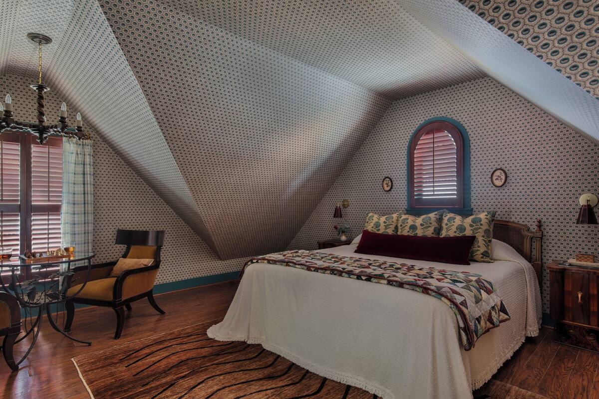 Une chambre avec un plafond en pente, un lit recouvert d’une couvre-lit à motifs, un plancher en bois et une table à manger avec des chaises.