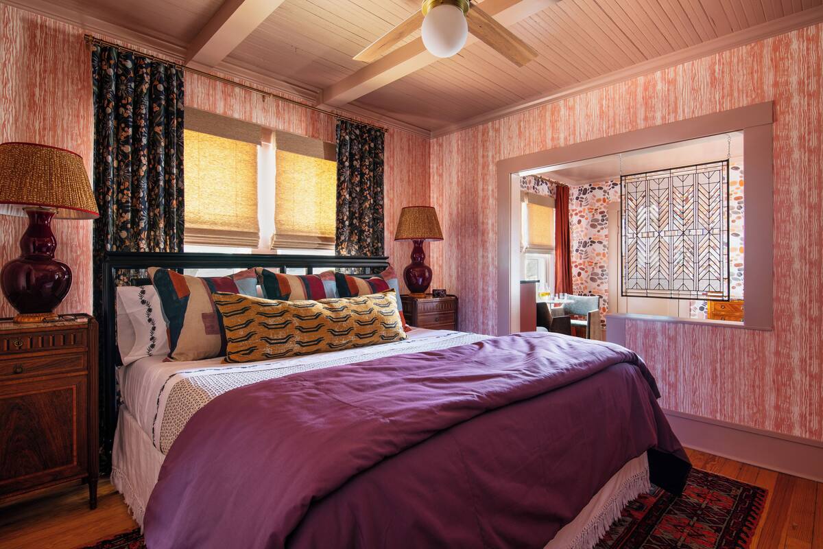 Une chambre avec un plafond en bois, un lit recouvert d’une courtepointe violette, des rideaux à motifs et une fenêtre offrant une vue décorative.