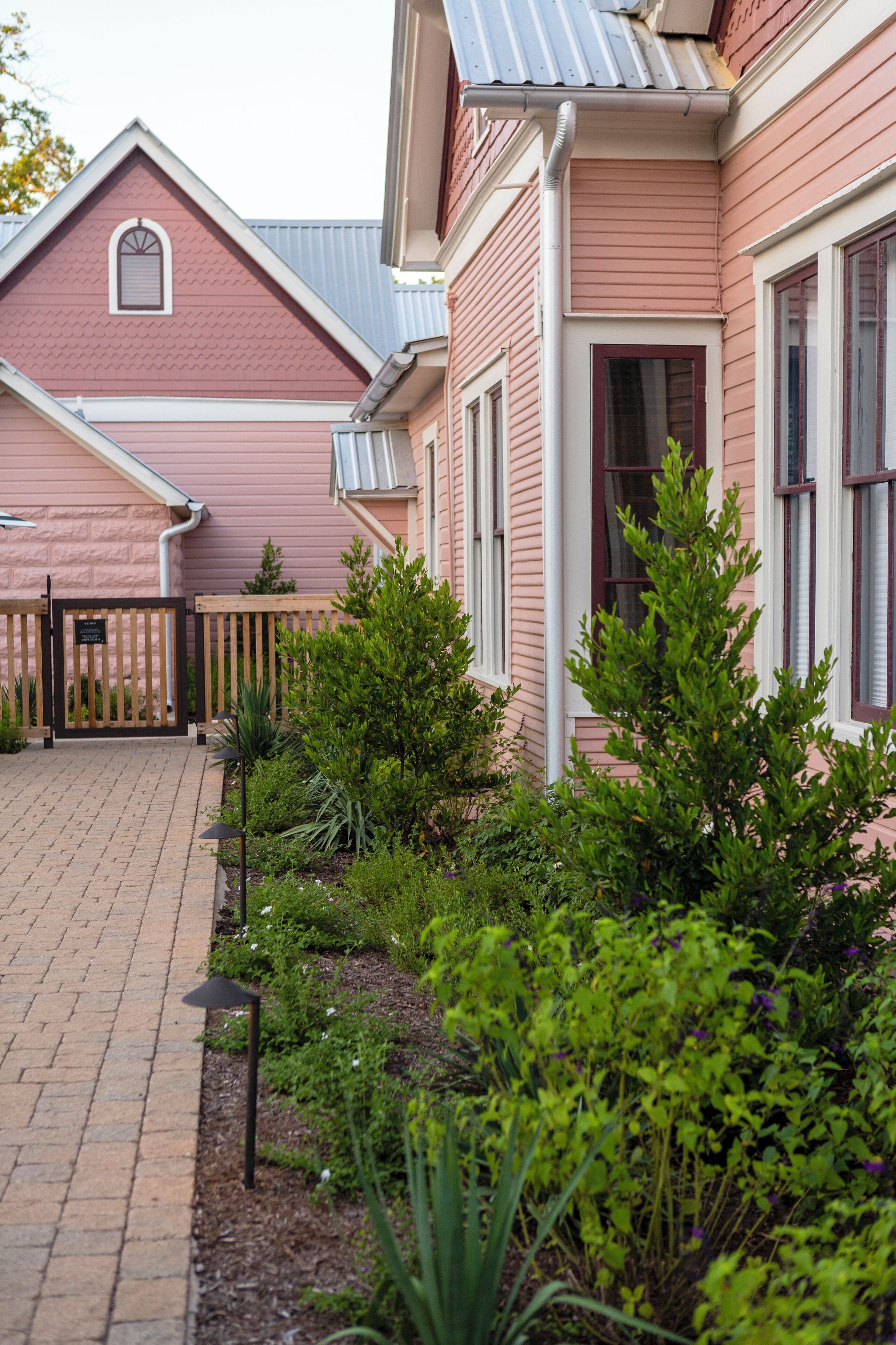 A pink building with a gated entrance, a brick pathway, and green shrubs.