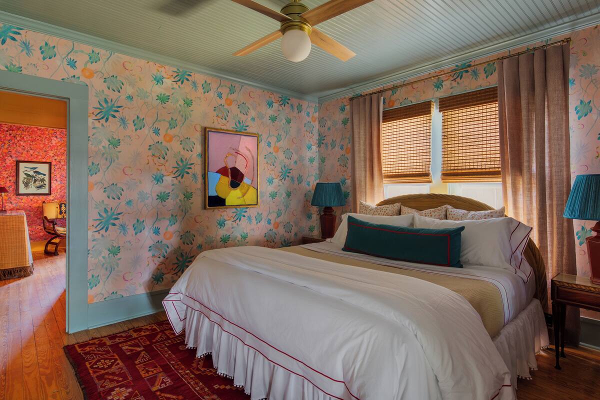 Une chambre avec un grand lit, un ventilateur de plafond, une fenêtre avec des stores et un mur décoré d’un motif.