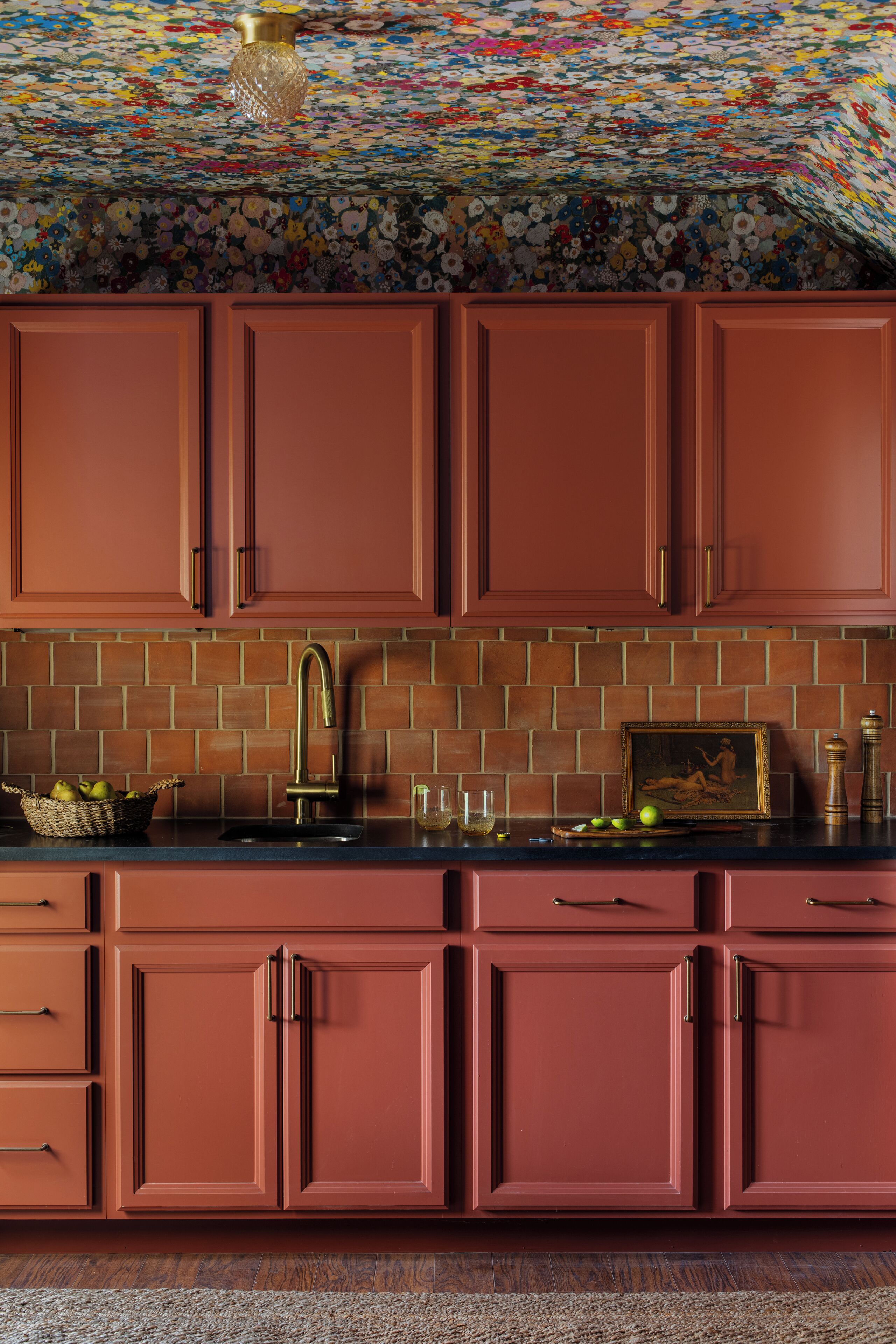A kitchen with red cabinets, a black countertop, a tiled backsplash, and a floral ceiling.