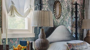 A room with a bed, a table with a lamp, a chair, and a window with curtains. - The Menagerie (Fredericksburg)