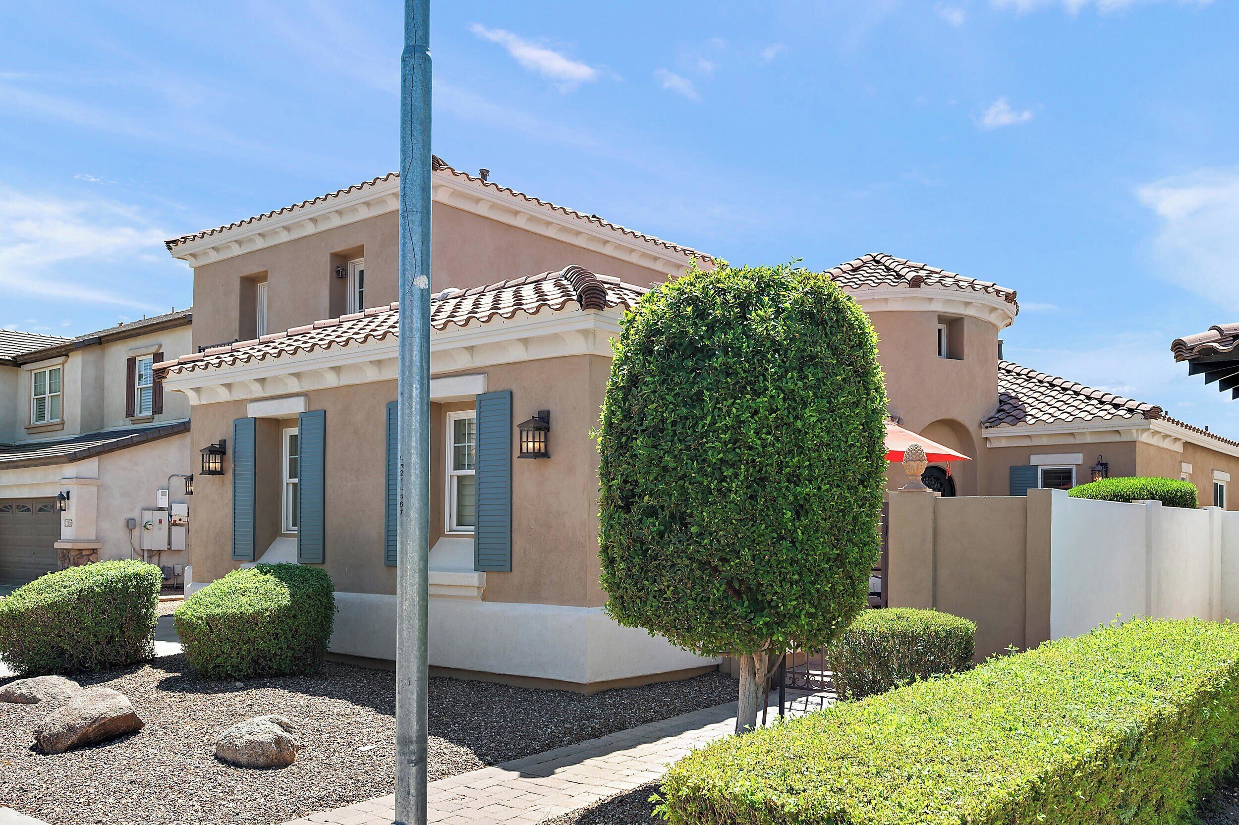 A two-story house with a tiled roof, a beige exterior, and a small front yard with trimmed bushes and rocks.