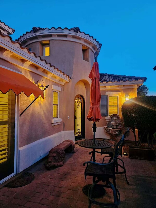 A two-story house with a patio area featuring a round table and chairs, a red umbrella, and a decorative fountain. - Chandler Keep – Medieval Spanish Villa, BBQ Courtyard & Prime South Chandler (Chandler)