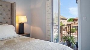 Ein Schlafzimmer mit Bett, Nachttisch, Lampe und einem Balkon mit Blick auf Gebäude und eine Pflanze.