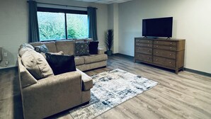 Ein Wohnzimmer mit einem beigefarbenen Sofa, einer Holzkommode, einem Flachbildfernseher und einem großen Fenster mit Vorhängen.