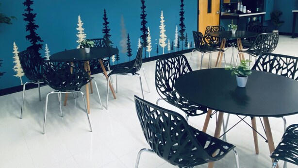 Ein Zimmer mit blauer Wand, die ein Waldgemälde zeigt, und der Aufschrift 'Tree Top Breakfast Bistro'. Mehrere schwarze Tische und Stühle sind zum Essen angerichtet.