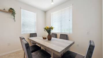 A dining area with a rectangular table, six chairs, a vase with flowers, and two windows with blinds.