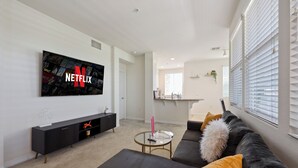 Una sala de estar moderna con un televisor de pantalla plana grande que muestra el logotipo de Netflix, un sofá oscuro con cojines amarillos y blancos, una mesita de centro redonda y un mueble de entretenimiento negro.