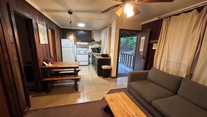 Ein gemütlicher Wohnbereich mit einer Couch, einer hölzernen Bank und einem Esstisch. Eine Deckenventilator und Pendelleuchten sind sichtbar. Im Hintergrund befindet sich eine Küche mit Kühlschrank, Herd und Mikrowelle.