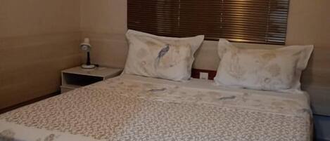 A bedroom with a bed, a nightstand, a wall-mounted air conditioner, and a window with blinds.