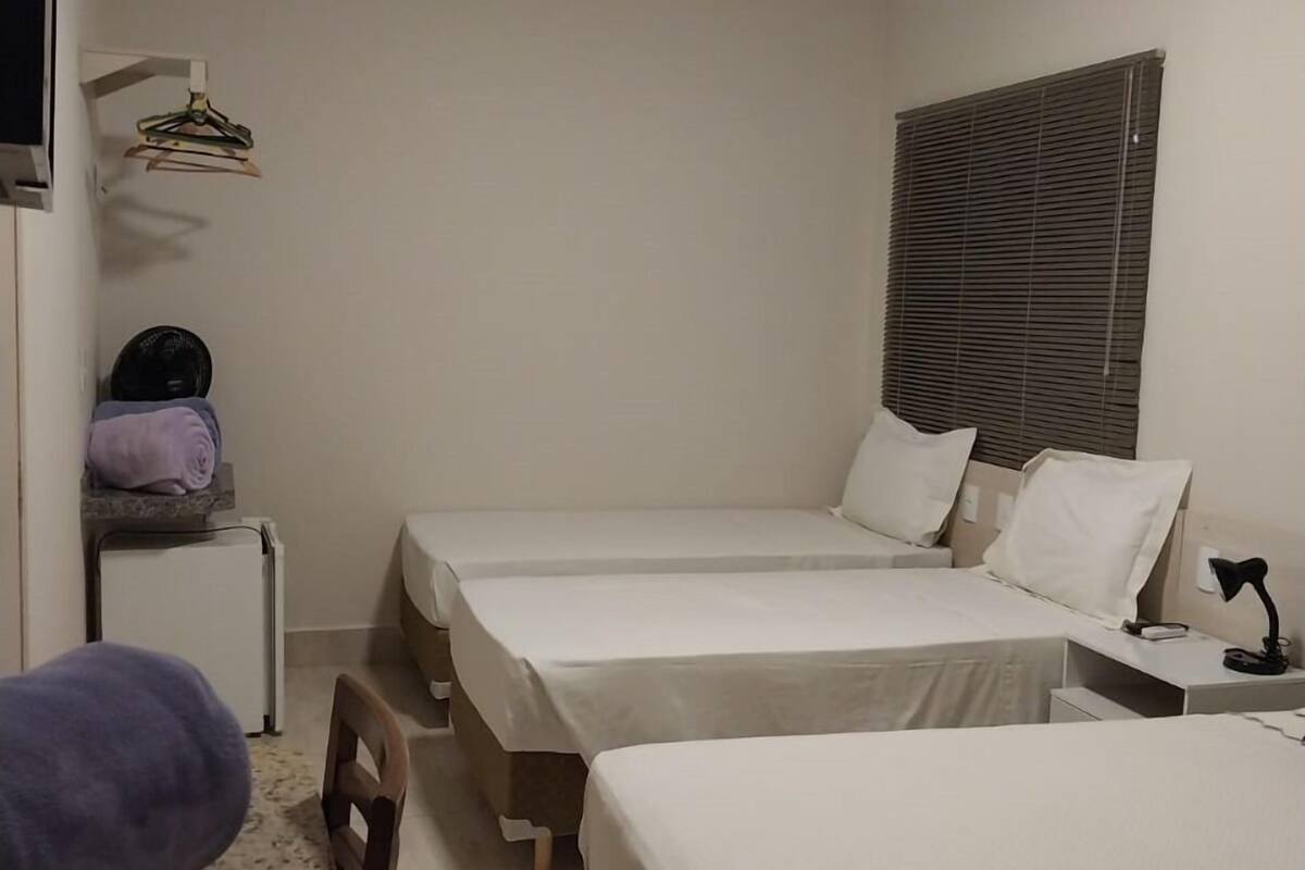 A small, clean room with two single beds, a desk, a chair, a small refrigerator, and a window with blinds.