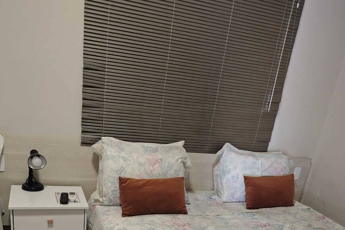 A bedroom with a bed, two pillows, a nightstand with a lamp, and a window with blinds.