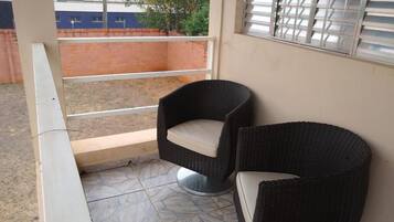 Un balcón con dos sillas de mimbre, piso de baldosas y vista a un edificio al fondo.