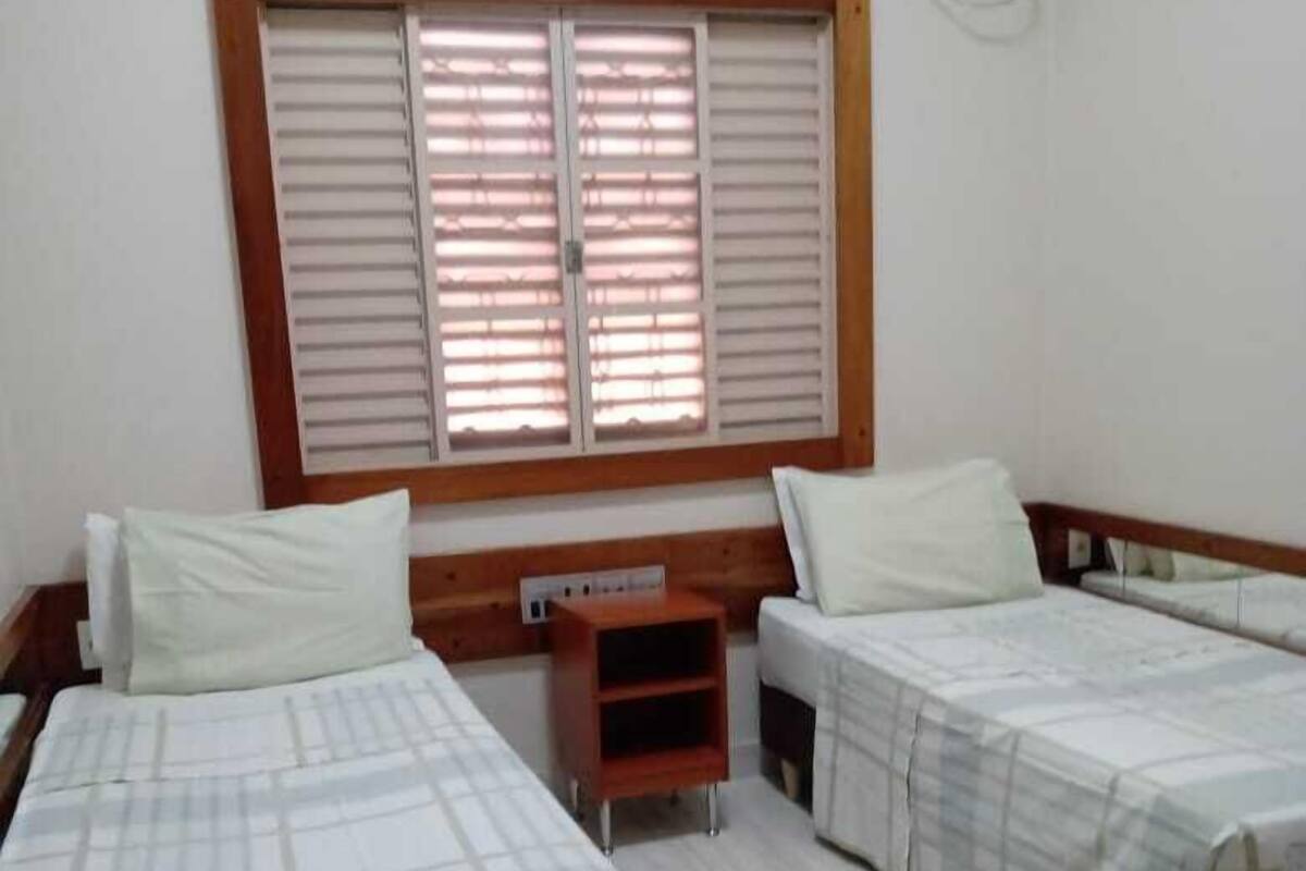 A room with two beds, a small wooden nightstand, a window with blinds, and a wall-mounted air conditioning unit.