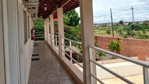 Un balcón techado con suelo de baldosas, barandillas blancas y vistas a una piscina y vegetación.