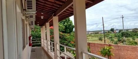 A covered balcony with a tiled floor, white railings, and a view of a swimming pool and greenery.