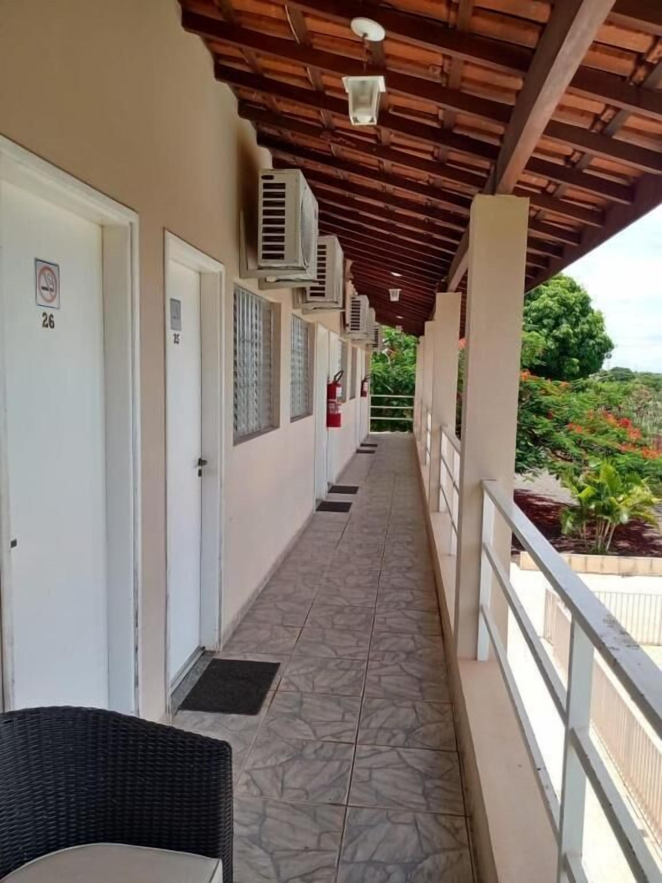 A covered corridor with doors numbered 26 and 27, air conditioning units, and a balcony with a view of greenery.