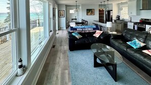 A spacious living room with large windows, a black leather sofa, a glass coffee table, and a kitchen area in the background. - The Place Up North (Traverse City)