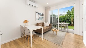 A modern living room with a white sofa, a dining table with chairs, and a large sliding glass door leading to a garden. - Coastal retreat near shops + beach (Corrimal)