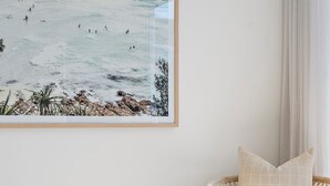 A room with a wicker chair, a framed artwork of a beach scene, and a woven rug. - Coastal retreat near shops + beach (Corrimal)