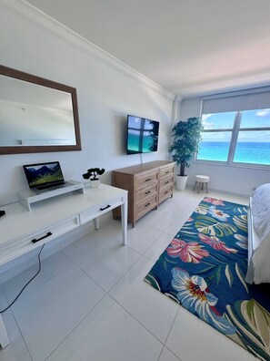 A modern bedroom with a large window, a flat-screen TV, a desk, a dresser, and a floral rug.