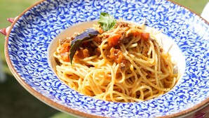 A bowl of spaghetti with a bay leaf garnish, held by a person with red nail polish, in front of a poolside setting with wooden chairs and a building in the background. - Q Alacati Otel (Cesme)